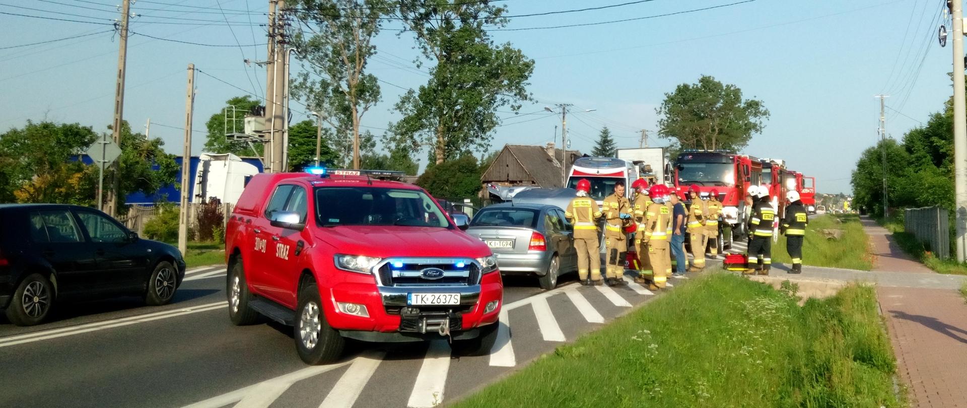 Na zdjęciu droga krajowa na której doszło do wypadku drogowego. Od lewej stoją: pierwsze uszkodzone auto (ciemne), samochód pożarniczy, kolejne uszkodzone auta (dwa jasne), fragment ambulansu za którym stoją: po prawej samochody pożarnicze a po lewej inni użytkownicy drogi. Na poboczu, na wysokości 2 uszkodzonych aut stoi grupa strażaków.