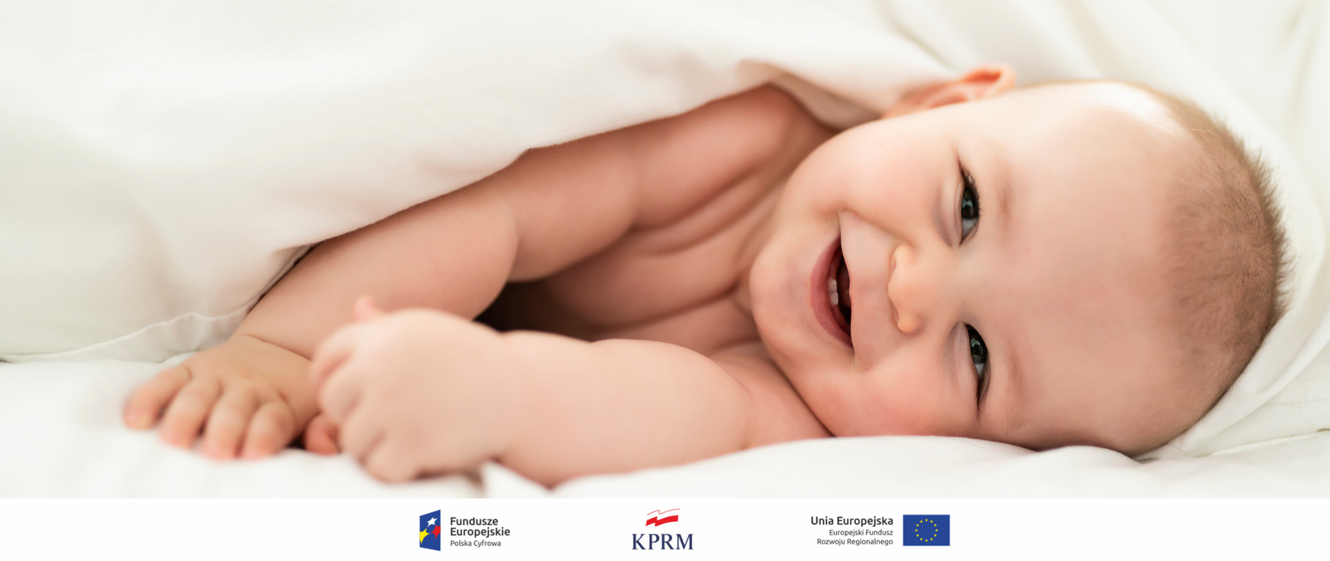 Leżący na boku, w białej pościeli, uśmiechnięty niemowlak.