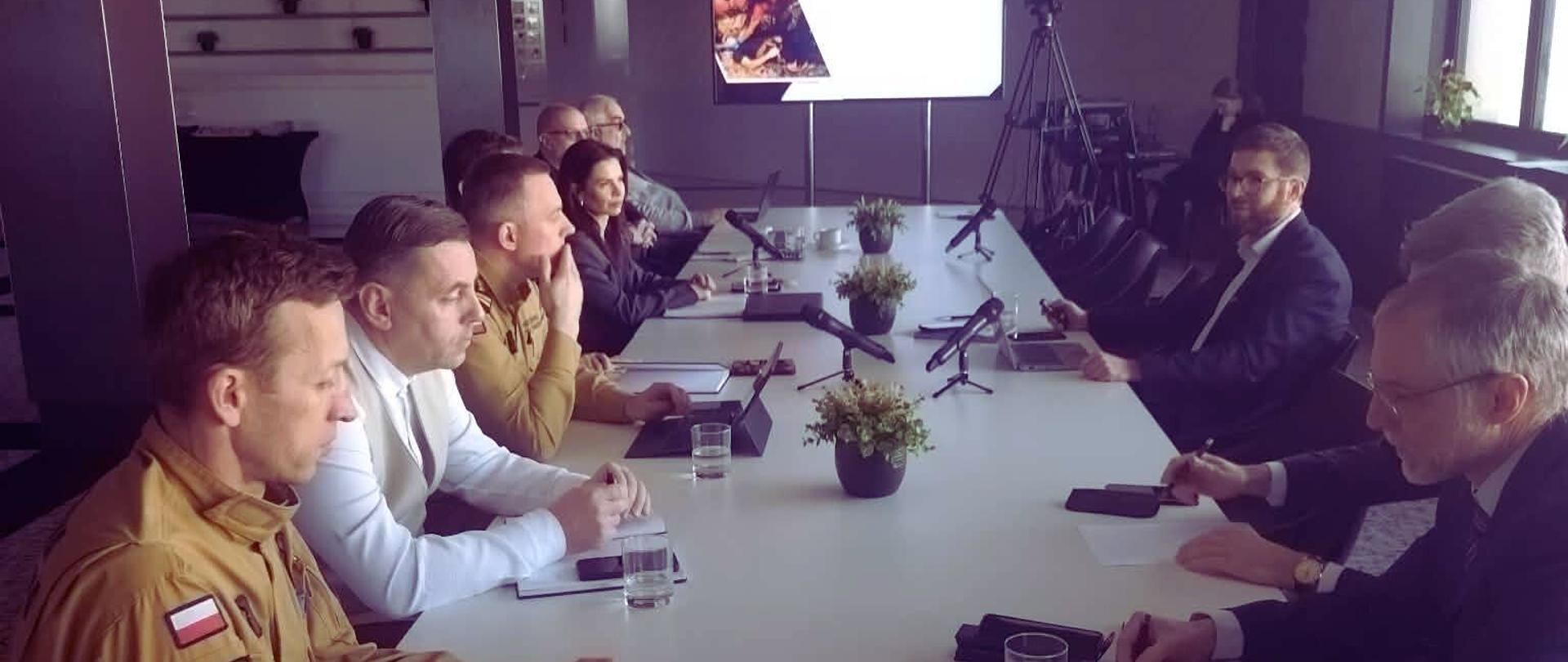 Członkowie rady programowej targów zasiadają przy wspólnym stole w sali konferencyjnej podczas spotkania poświęconemu tematyce wydarzenia. W tle znajduje się duży telewizor na stelażu, na którym wyświetlana jest prezentacja dotycząca wydarzenia.