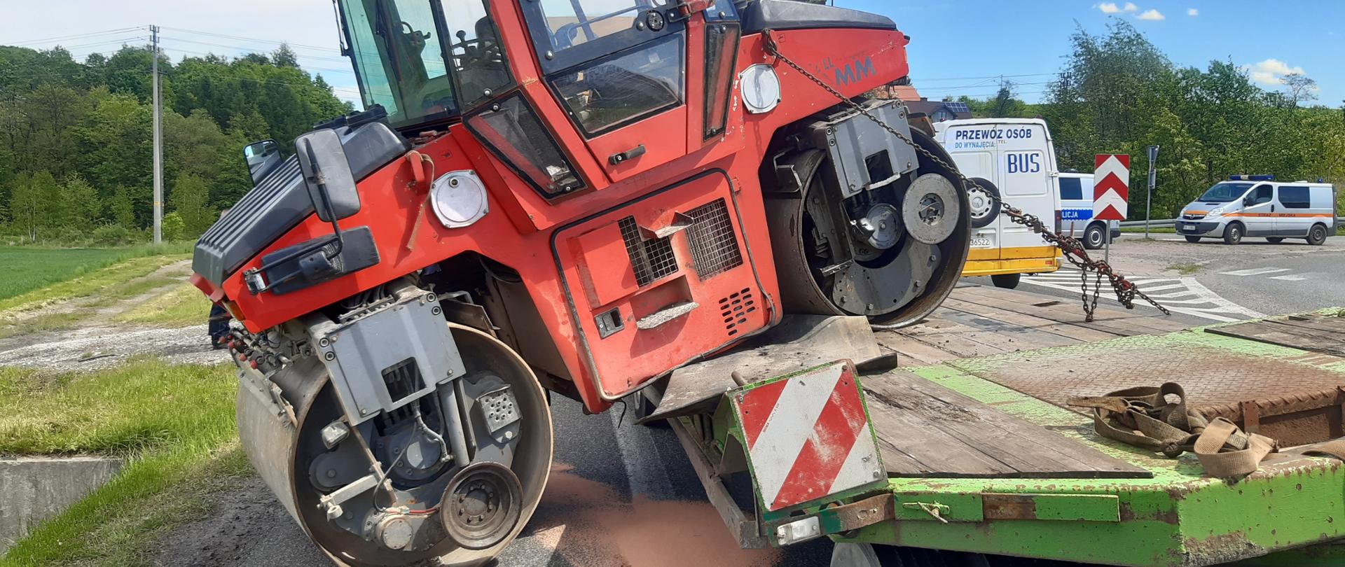 Na zdjęciu widoczny walec drogowy który częściowo stoi na naczepie ( przedni wał), a częściowo na drodze ( tylni wał). Walec znajduje się w poprzek naczepy, blokując jeden pas ruchu.