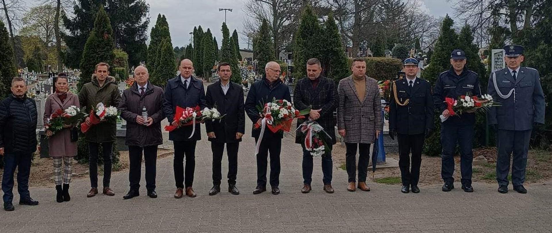 Dzień Pamięci Ofiar Zbrodni Katyńskiej. Cmentarz parafialny w Rawiczu. Delegacja rawickich służb mundurowych oraz władz samorządowych przed ceremonią składania wiązanek kwiatów. Wszyscy stoją w jednym szeregu. 