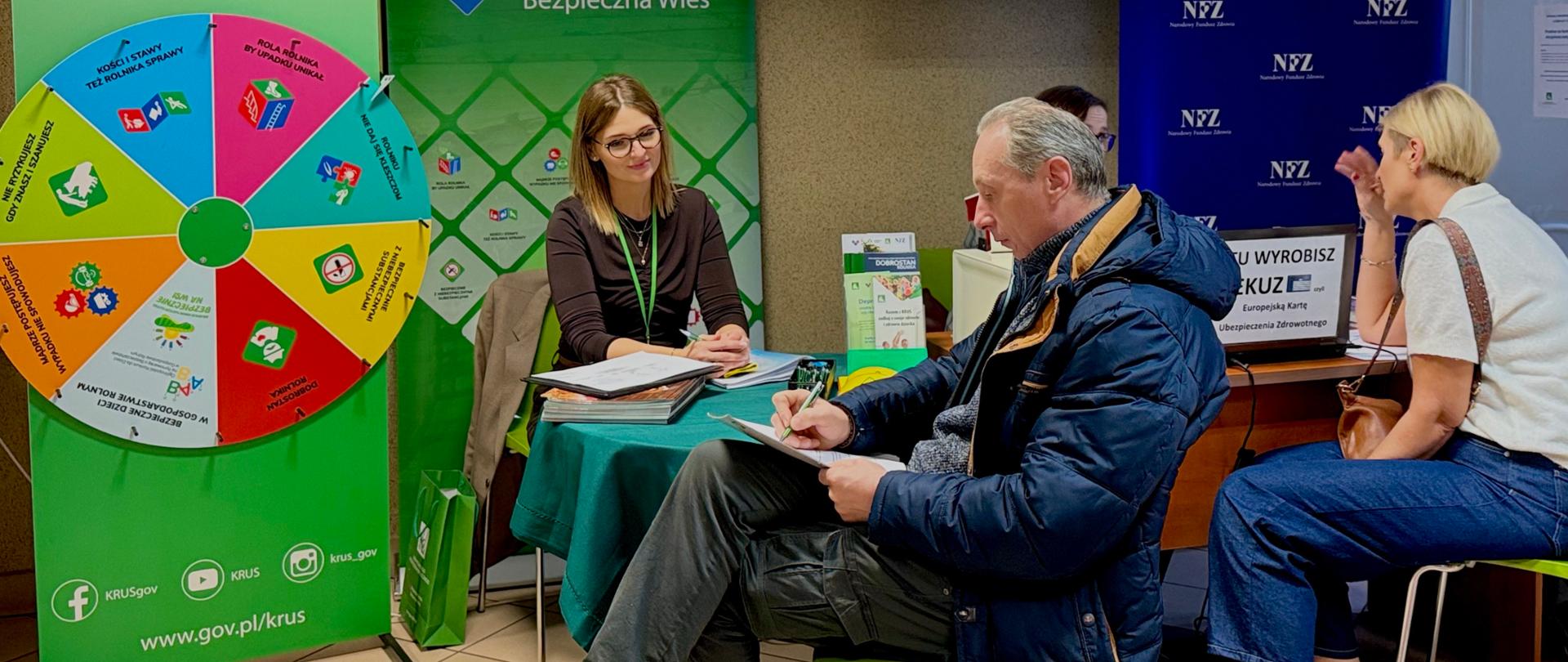 Przy stole informacyjnym siedzą przedstawicielki KRUS i NFZ. Kobieta z zielonym identyfikatorem rozmawia z uczestnikiem wydarzenia. W tle widoczne są banery KRUS i NFZ oraz kolorowe koło konkursowe „Bezpieczny Rolnik Bezpieczna Wieś”.
