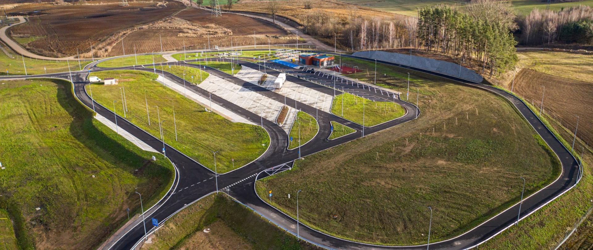 Widok z lotu ptaka na miejsca parkingowe dla aut osobowych i ciężarowych. W oddali pola użytkowe.