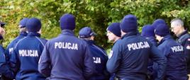 Grupa policjantów w granatowych mundurach i czapkach zimowych stoi tyłem i bokiem do obiektywu, tworząc krąg podczas odprawy. Na plecach ich bluz widoczne są duże białe napisy „POLICJA”. Po prawej stronie w tle widać również strażaka.