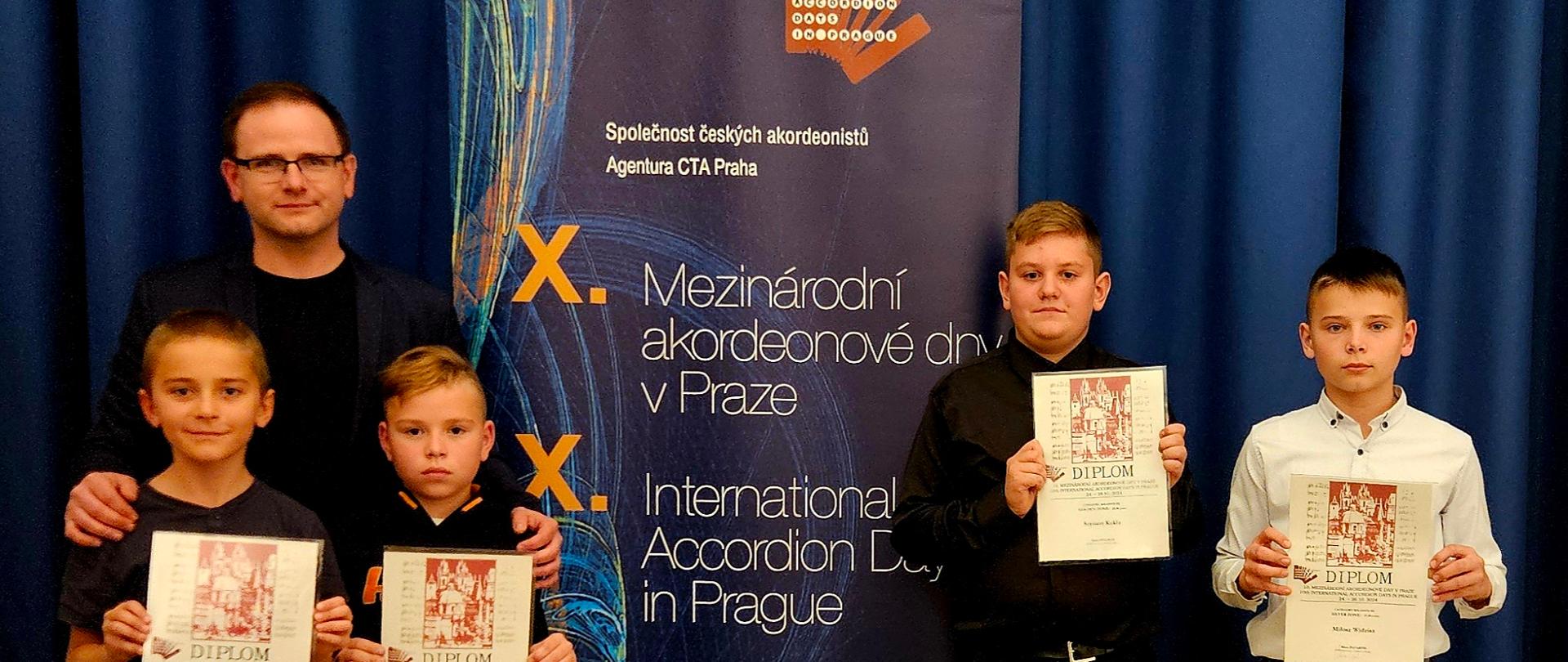 Stojący obok rollup-a uczniowie prezentujący zdobyte dyplomy wraz z nauczycielem. Za ich plecami granatowa kotara