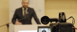 Minister funduszy i polityki regionalnej Grzegorz Puda podczas 5. Forum Akademicko-Gospodarczego, minister stoi przy mikrofonie i przemawia, na pierwszym planie kamera z podglądem obrazu na którym widąć ministra