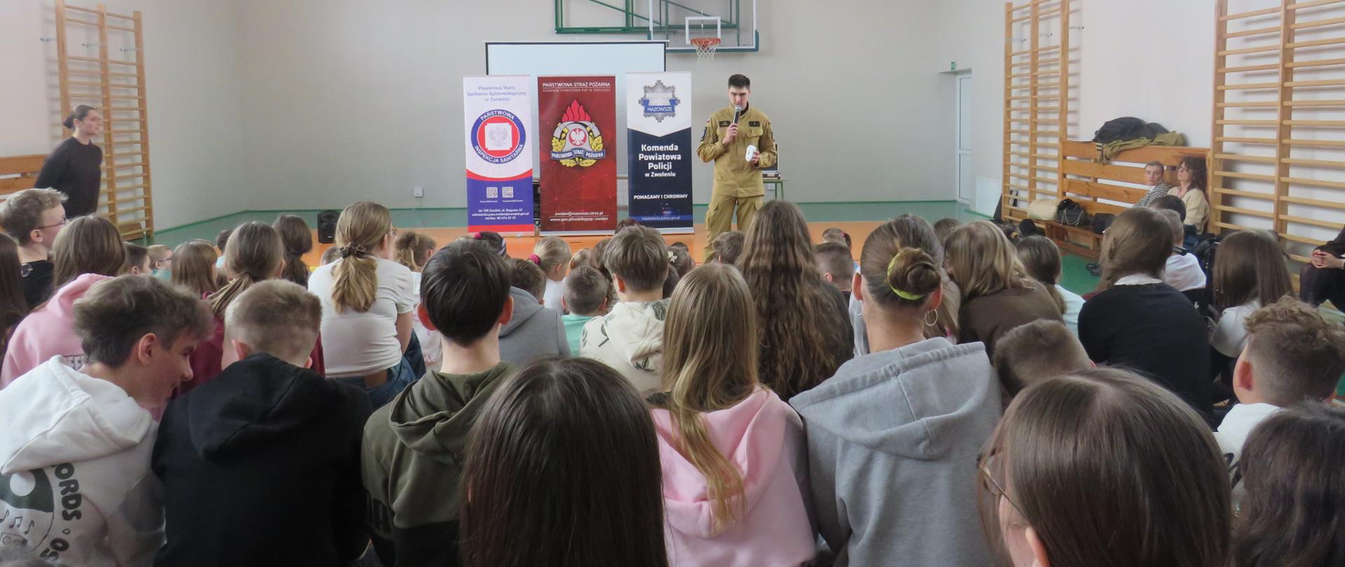 Spotkanie profilaktyczne w sali gimnastycznej – strażak przemawia do dużej grupy dzieci siedzących na ławkach, za nim widoczne banery służb oraz ekran projekcyjny.