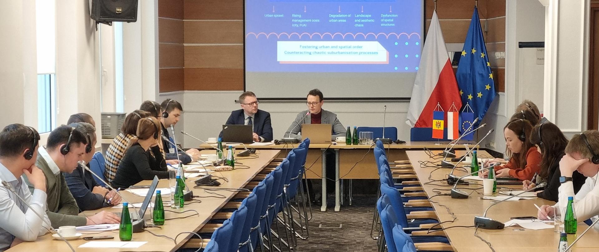 Sala konferencyjna ze stołami przy których siedzą ludzie z laptopami i słuchawkami. Wyświetlana jest prezentacja po angielsku, widać flagi Polski, Unii Europejskiej i Mołdawii