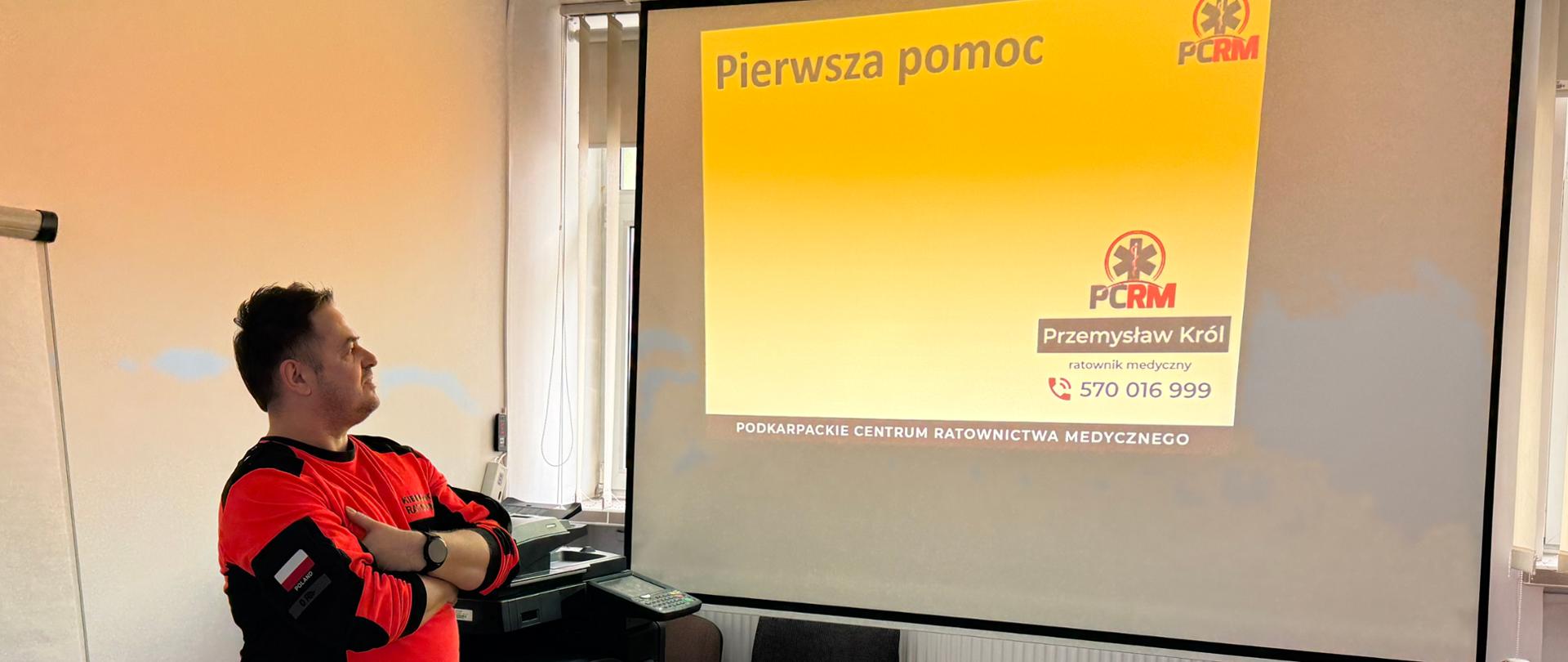 Mężczyzna w pomarańczowo czarnej koszulce stoi przed wyświetlaną prezentacją.
