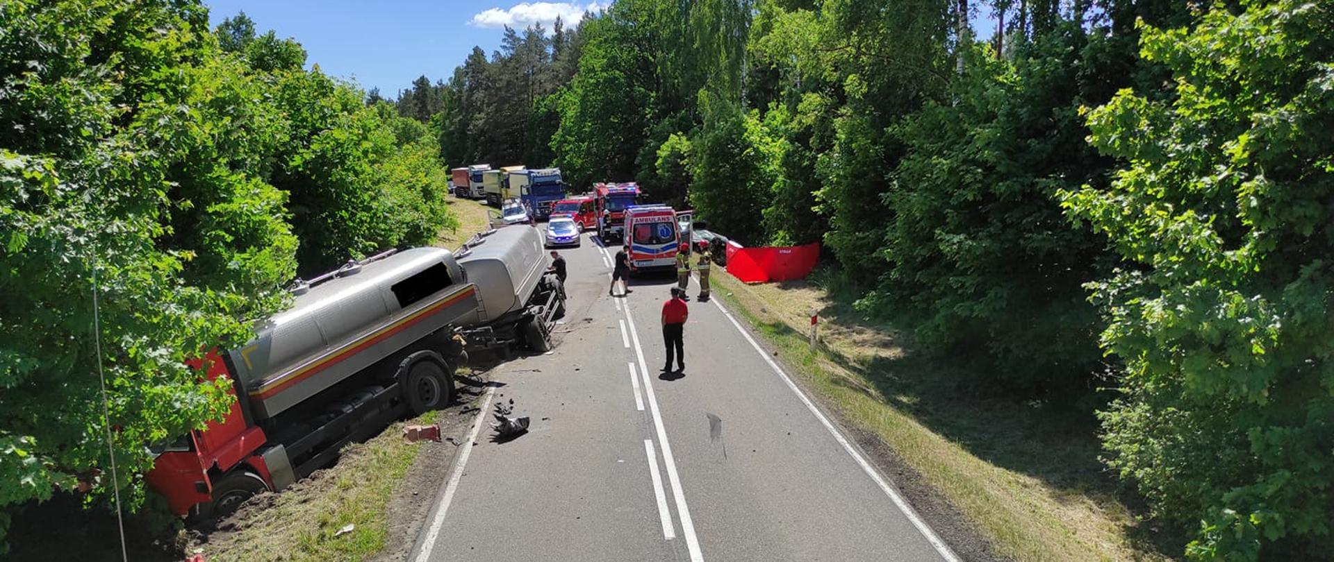 Zdjęcie przedstawia miejsca wypadku na drodze DK 57, na wysokości miejscowości Kucbork. Samochód ciężarowy częściowo w rowie częściowo na jezdni po lewej stronie, samochody osobowe po prawej stronie jezdni w rowie. Na miejsce zastępy straży pożarnej, zespół ratownictwa medycznego oraz patrol policji.