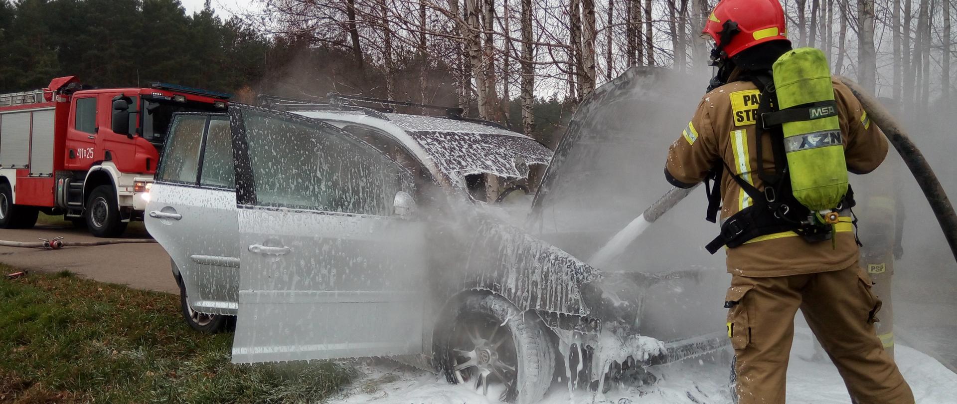 Strażak podczas gaszenia komory silnika samochodu osobowego. Przód samochodu oraz teren pod samochodem pokryty pianą gaśniczą. W tle samochód strażacki.