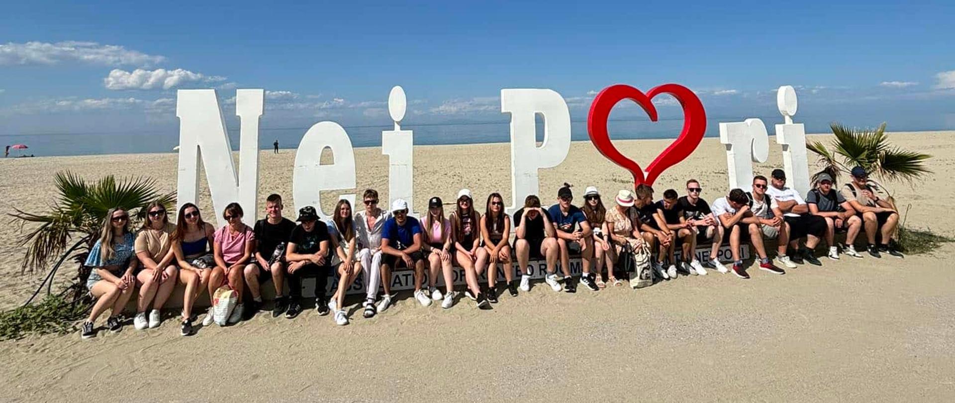 Duża grupa osób siedzi na piasku przed wielkim, białym napisem „Nei Pori” z czerwonym sercem zamiast litery „o”. Zdjęcie wykonane na szerokiej, pustej plaży pod bezchmurnym niebem.