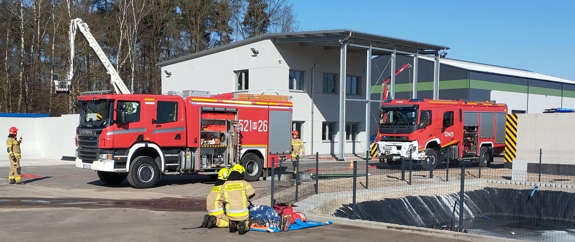 Ćwiczenia ratownicze PSP Mława na terenie suszarni zbóż – strażacy prowadzą symulowaną akcję ratowniczą z udzielaniem pomocy poszkodowanemu, a w tle stoją dwa wozy strażackie i pracuje podnośnik.