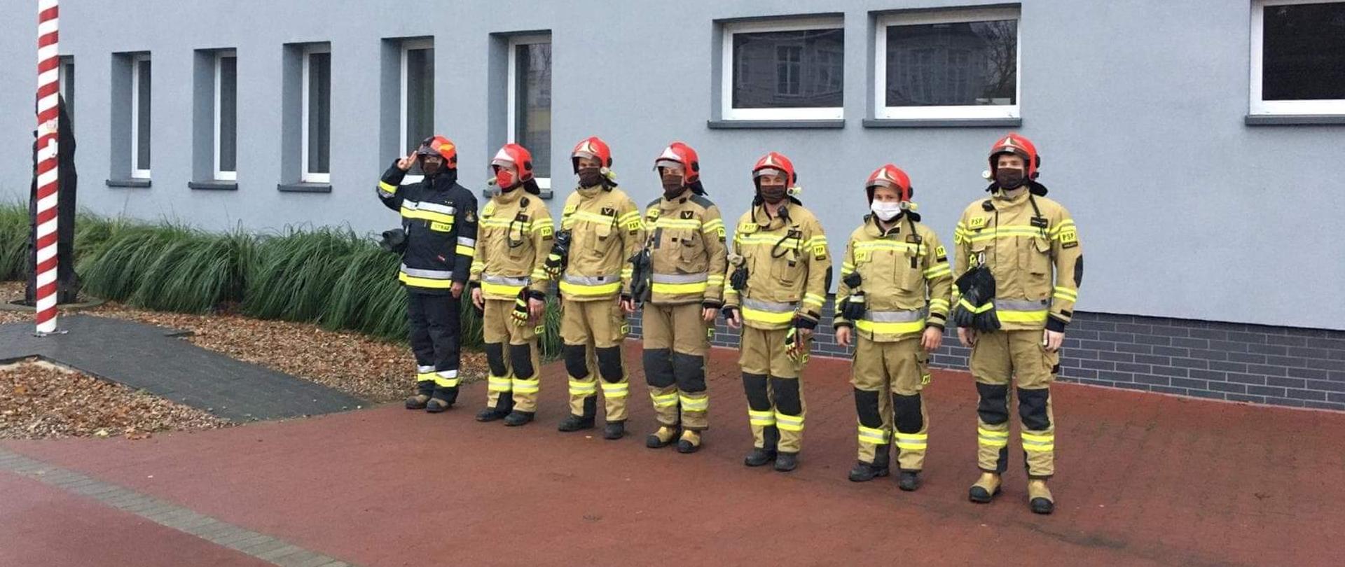 Siedmiu strażaków stoi przed budynkiem komendy. Dowódca oddaje honor. 
