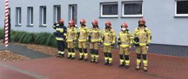 Siedmiu strażaków stoi przed budynkiem komendy. Dowódca oddaje honor. 
