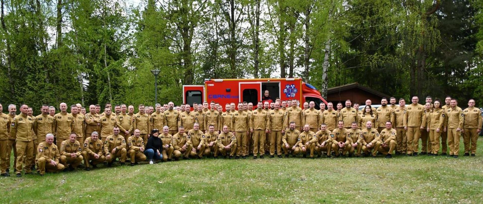 Grupowe zdjęcie uczestników narady na tle samochodu ratownictwa chemicznego do wykrywania skażeń pierwszy szereg kuca drugi szereg stoi.W tle widać drzewa