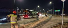 Strażacy z miotłami usuwają plamę oleju z drogi w tle autobus miejski oraz samochód osobowy.