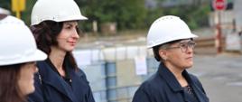Wiceminister Mrówczyńska w białym kasku stoi na dworze, obok niej dwie kobiety w takich samych kaskach, w tle palety z białymi zbiornikami.