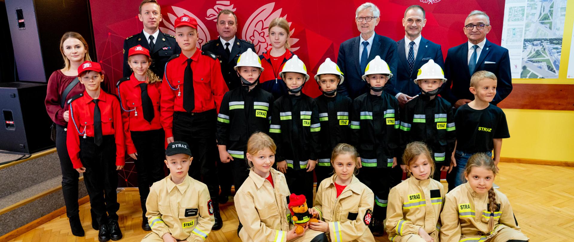 Zdjęcie przedstawia młodzieżowe drużyny pożarnicze, komendantów, posła oraz wójta stojących na tle komendy podczas uroczystego wręczenia prosem 
