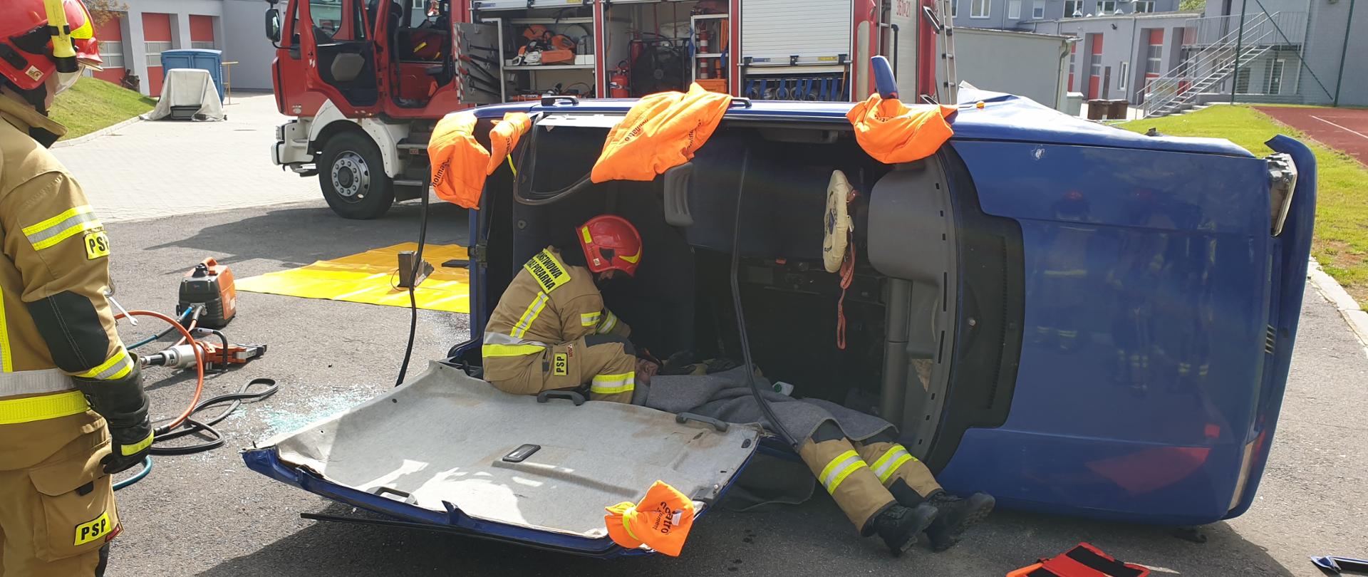 Zdjęcie przedstawia strażaków podczas ćwiczeń na placu jednostki. Samochód osobowy leży na boku, ma odcięty dach i następuje ewakuacja poszkodowanego. Na zdjęciu widać również samochód strażacki 