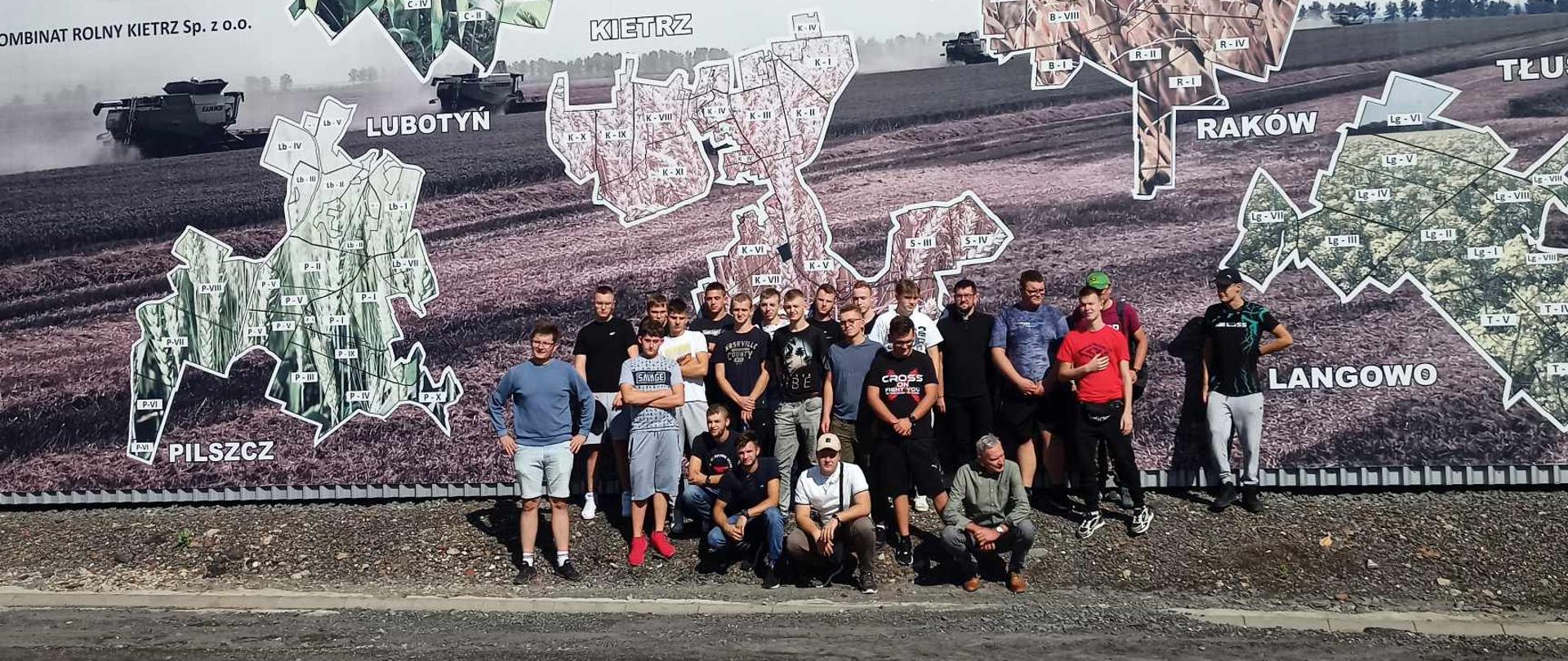 Grupa uczniów i opiekunów pozuje do pamiątkowego zdjęcia na tle wielkoformatowej mapy pól uprawnych Kombinatu Rolnego Kietrz. Mapa przedstawia różne kompleksy rolne, takie jak Lubotyń czy Pilszcz.