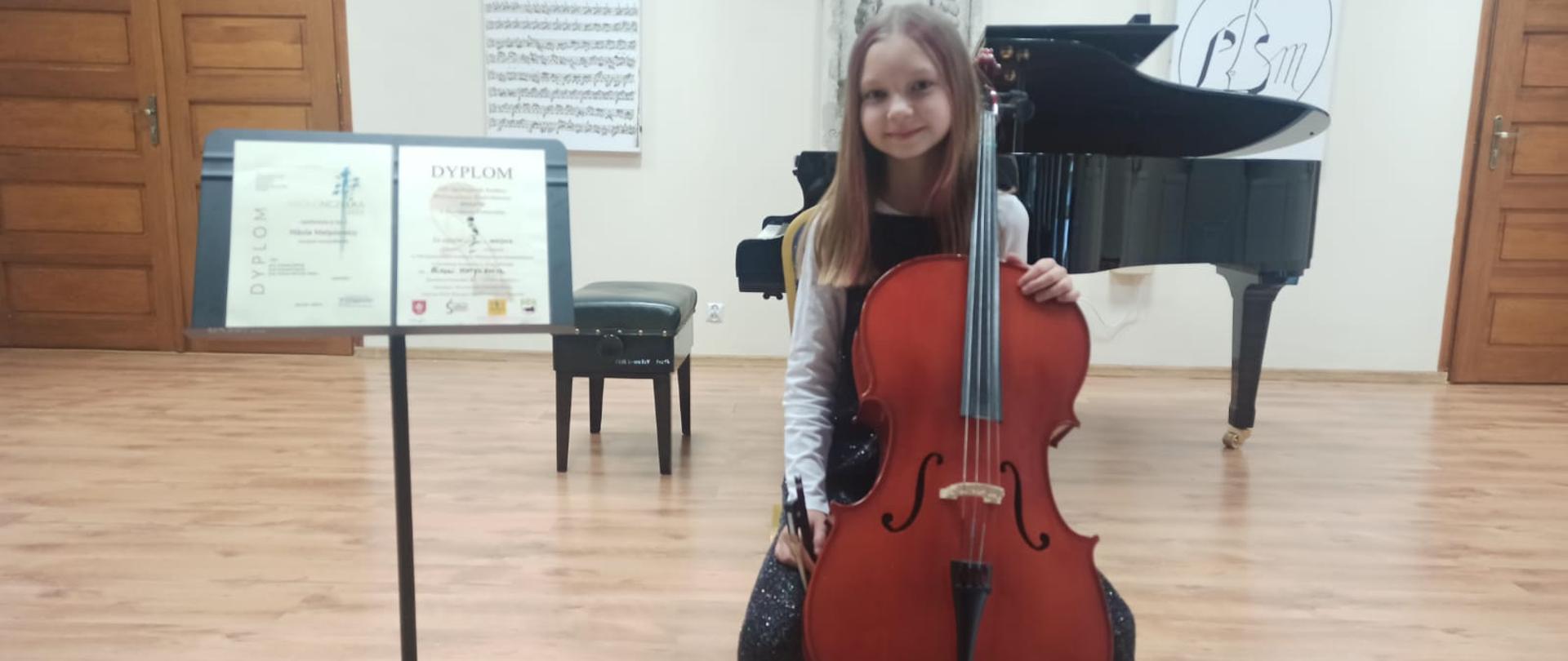 Na zdjęciu siedzi dziewczynka trzymając wiolonczelę obok ustawiono pulpit z Dyplomami w tle z napisami Dyplom Wyróżnienia i Dyplom za zajęcie II miejsca.. W tle fortepian i portrety Jana Sebastiana Bacha, oraz Logo Szkoły i partytura J. S. Bacha.