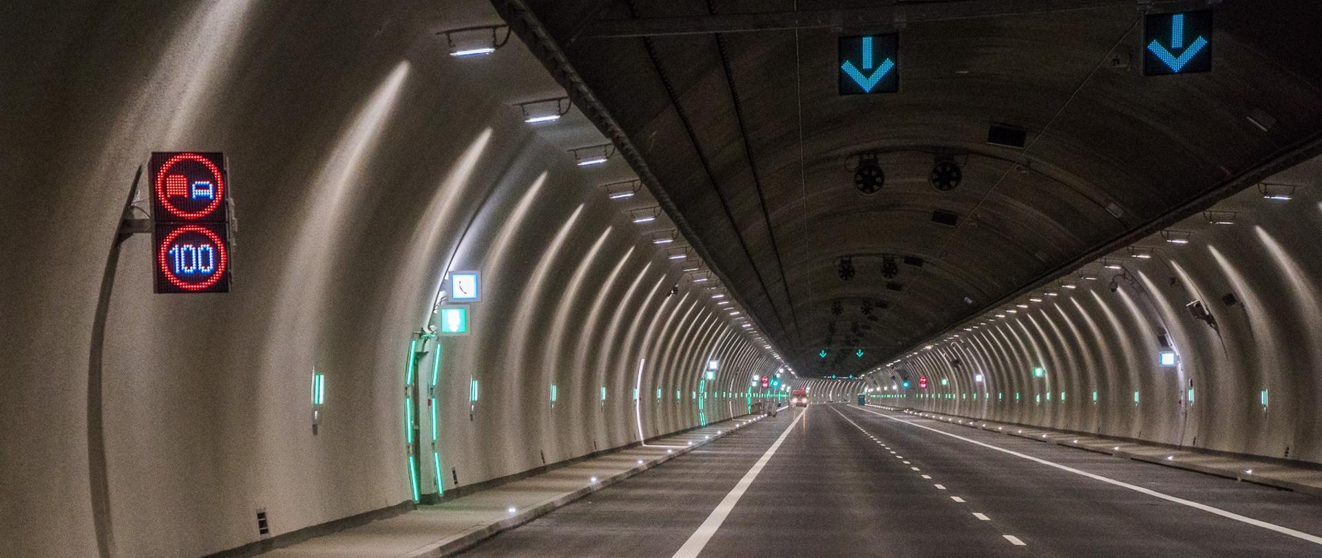 zdjęcie przedstawia wnętrze tunelu z widocznym oznakowaniem poziomym, widocznym oznakowaniem ograniczenia prędkości i zakazu wyprzedania i strzałkami wskazującymi na wolny pas.