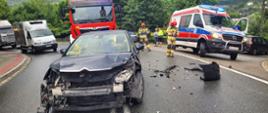 Samochód z uszkodzonym przodem na drodze. W tle samochód pożarniczy, karetka Pogotowia ratunkowego,inne pojazdy oraz strażacy.