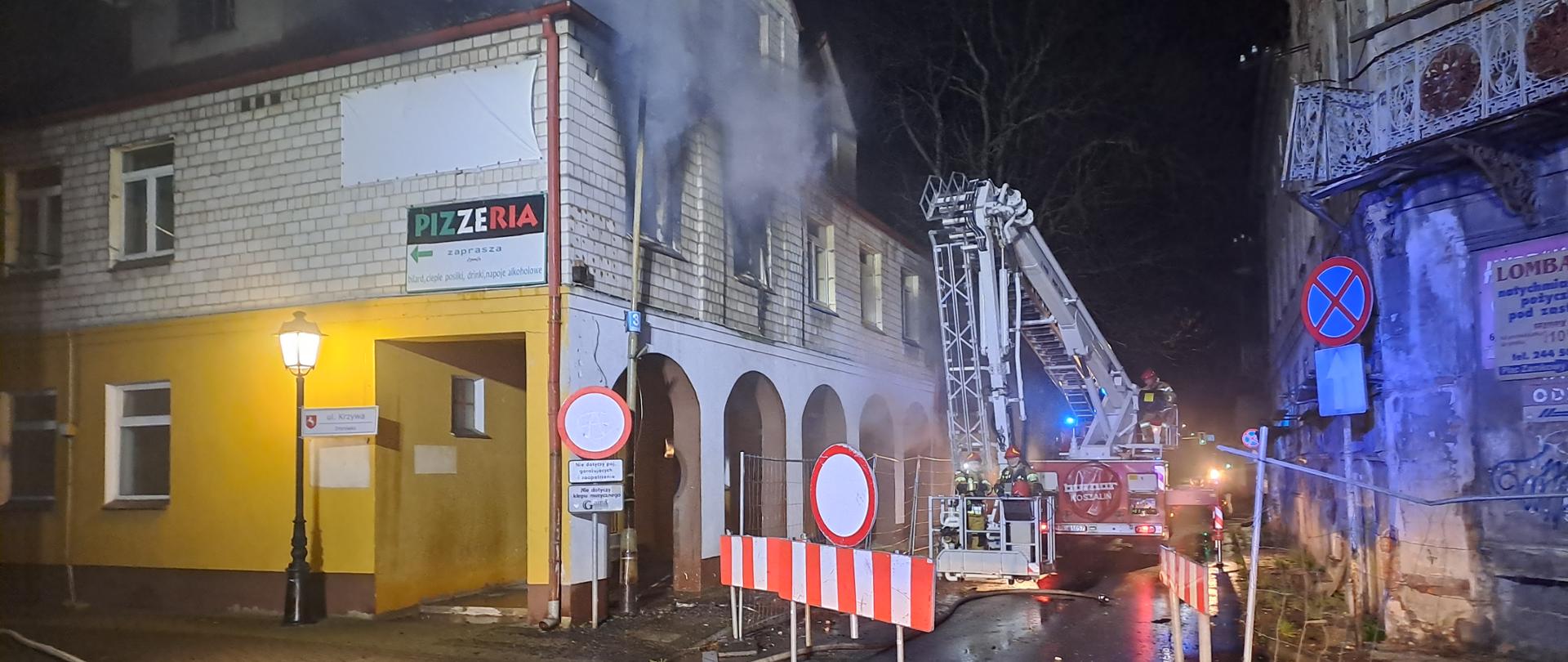 Zdjęcie przedstawia pożar kamienicy. Z okien na piętrze wydobywał się dym. Przy budynku stoi drabina strażacka, w koszu drabiny stoją strażacy. Przed budynkiem widać linie wężowe. 