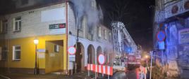 Zdjęcie przedstawia pożar kamienicy. Z okien na piętrze wydobywał się dym. Przy budynku stoi drabina strażacka, w koszu drabiny stoją strażacy. Przed budynkiem widać linie wężowe. 