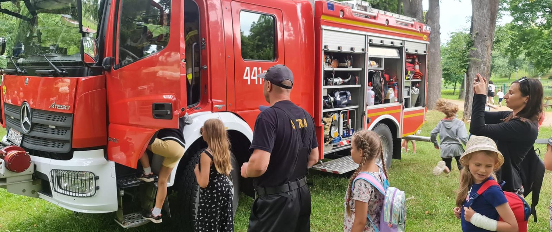 Obraz przedstawia strażaka pokazującego dzieciom samochód pożarniczy