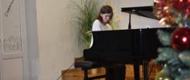Na zdjęciu znajduje się uczennica grająca na czarnym fortepianie, po prawej stronie fragment choinki oraz szkolnego logo na ścianie. Podłoga-scena drewniana, kolor ścian jasny