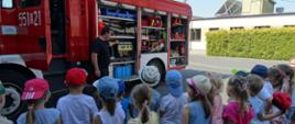 Zdjęcie przedstawia przedszkolaków słuchających strażaka podczas pogadanki na tle samochodu bojowego z otwartymi skrytkami bocznymi i widocznym sprzętem