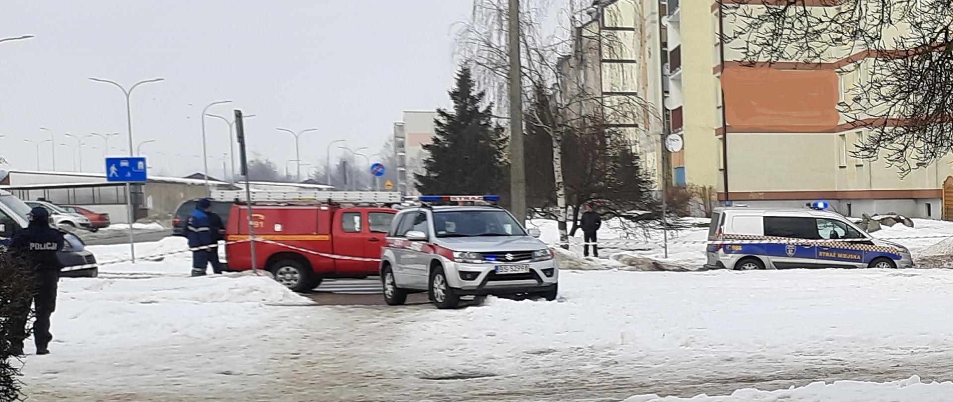 Zdjęcie obrazuje osobowy samochód strażacki osobowy samochód straży miejskiej samochód pogotowia energetycznego w lewej stronie zdjęcia stoi policjant w tle kilka osób i budynki. Aura zimowa.