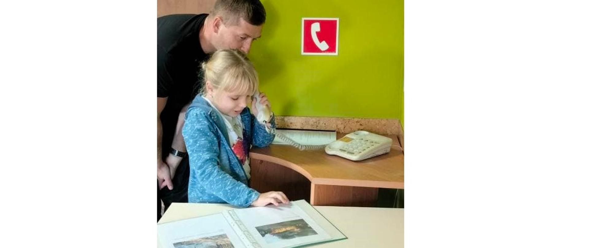 Dziewczynka dzwoni telefonem stacjonarnym, obok stoi strażak