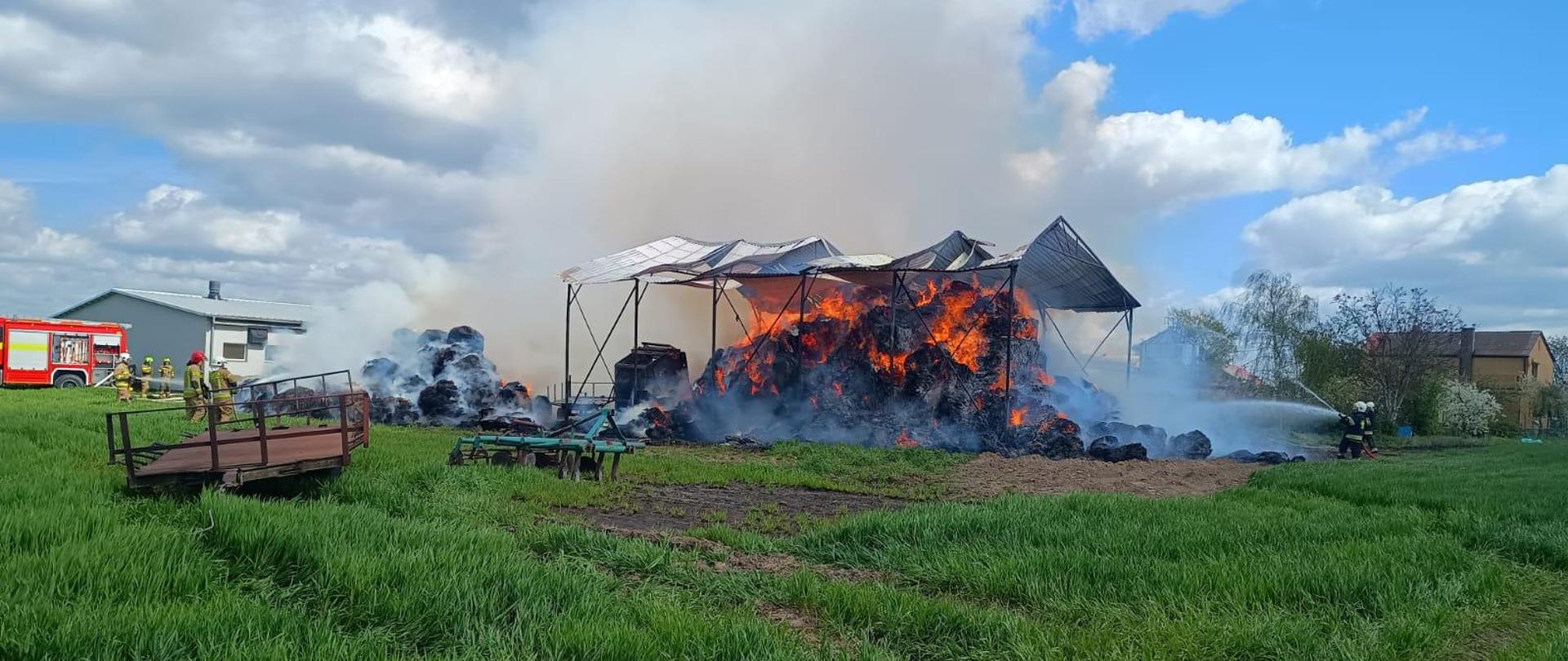 Na zdjęciu widać stalową wiatę pod którą palą się baloty słomy. Blaszany dach wiaty odkształcił się w wyniku oddziaływania termicznego. Strażacy tłumią ogień przy użyciu prądów gaśniczych.