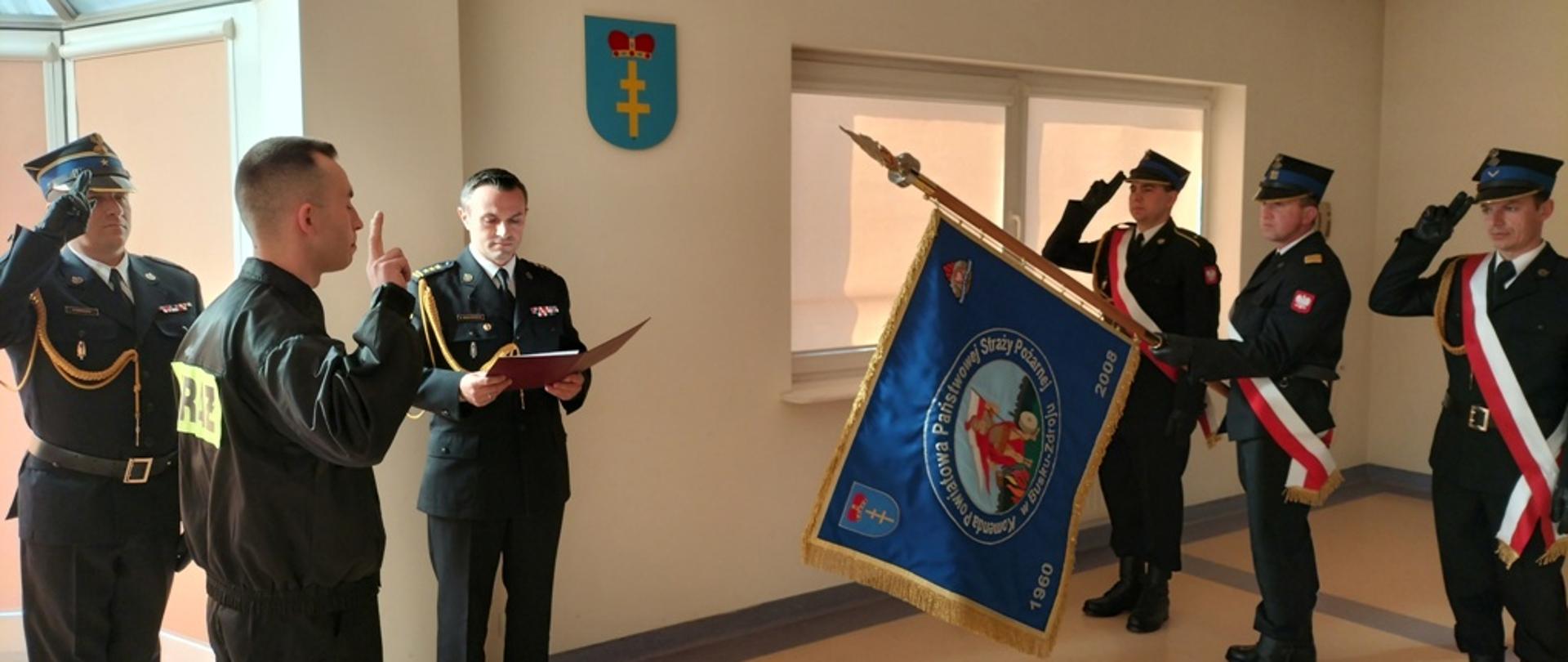 Zdjęcie przedstawia trzech strażaków w poczcie sztandarowym oddający honor przed którymi stoi nowo przyjęty do służby strażak składający ślubowanie z podniesioną prawą ręką. W tle komendant powiatowy odczytujący słowa roty ślubowania oraz dowódca uroczystości salutujący. 