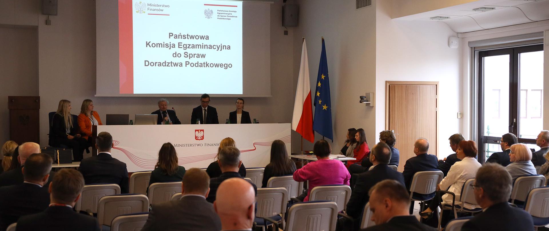 Prowadzący i uczestnicy spotkania, w tle napis Państwowa Komisja Egzaminacyjna ds. Doradztwa Podatkowego