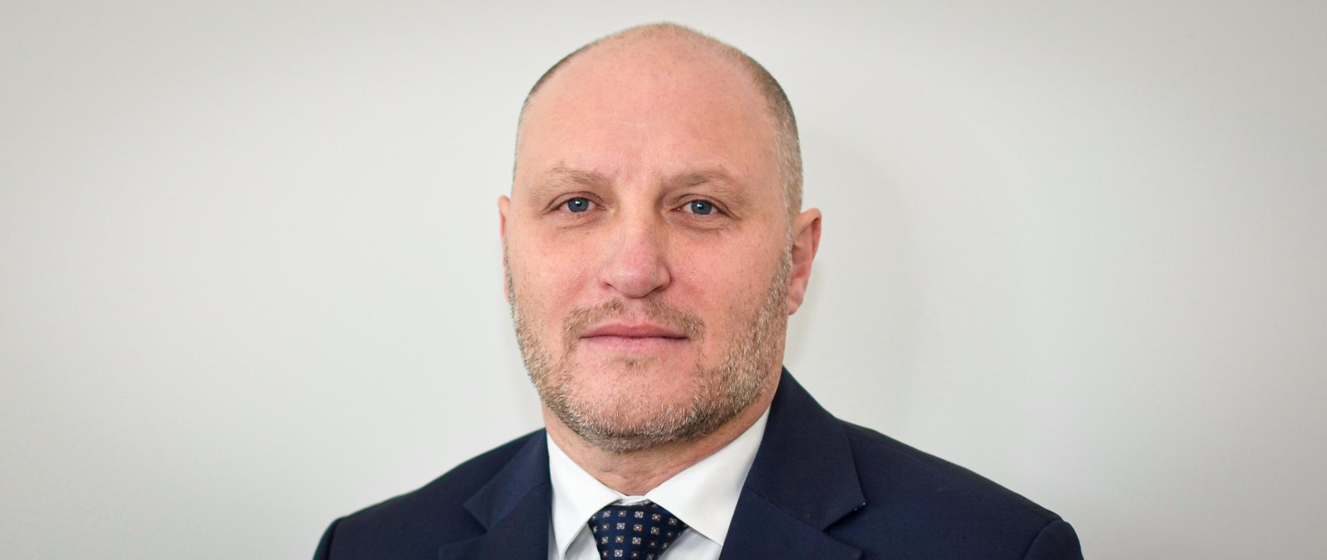 Mężczyzna w ciemnej marynarce, białej koszuli, ciemnym krawacie we wzorek, z krótkim zarostem