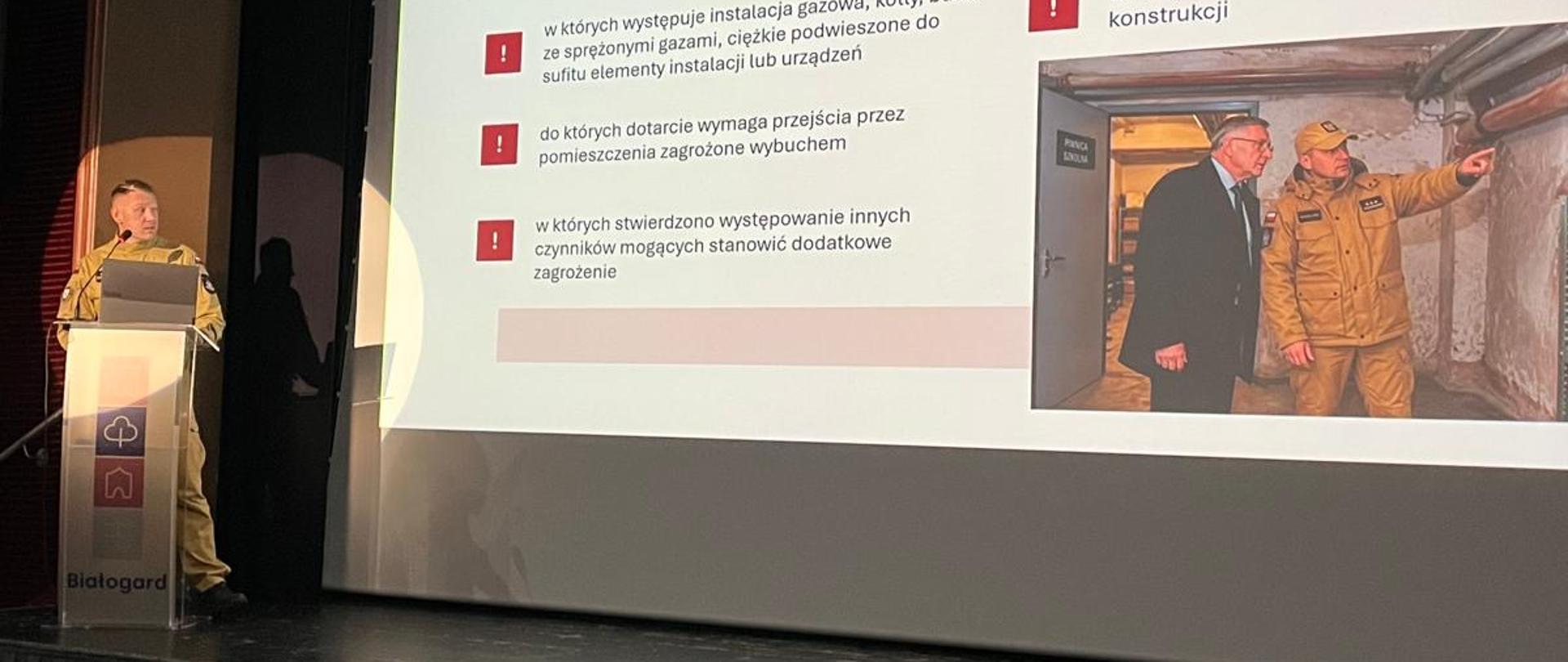 Strażak w piaskowym mundurze służbowym prowadzi prelekcję na scenie sali widowiskowej. Za nim znajduje się duży ekran multimedialny z prezentacją zatytułowaną: „Przestrzenie, których nie należy wskazywać na miejsce schronienia”. Na slajdzie wyliczone są czynniki ryzyka (m.in. duże przeszklenia, instalacje gazowe, zagrożenie wybuchem czy uszkodzenia konstrukcji) oraz umieszczone jest zdjęcie dwóch mężczyzn (w tym strażaka) dokonujących przeglądu piwnicy. Na mównicy widnieje logo Białogardu.