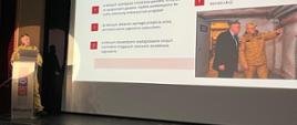 Strażak w piaskowym mundurze służbowym prowadzi prelekcję na scenie sali widowiskowej. Za nim znajduje się duży ekran multimedialny z prezentacją zatytułowaną: „Przestrzenie, których nie należy wskazywać na miejsce schronienia”. Na slajdzie wyliczone są czynniki ryzyka (m.in. duże przeszklenia, instalacje gazowe, zagrożenie wybuchem czy uszkodzenia konstrukcji) oraz umieszczone jest zdjęcie dwóch mężczyzn (w tym strażaka) dokonujących przeglądu piwnicy. Na mównicy widnieje logo Białogardu.
