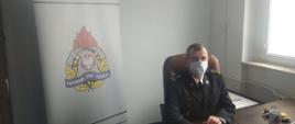 Komendant Miejski PSP w Bydgoszczy st. bryg. Janisław Buller siedzący przy biurku, ubrany w mundur wyjściowy, na twarzy maseczka. Na biurku laptop, telefon komórkowy. W tle za Komendantem ścianka z logo PSP i napisem "Komenda Miejska Państwowej Straży Pożarnej w Bydgoszczy"