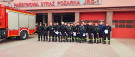 Na zdjęciu widzimy strażaków stojących w dwuszeregu. Strażacy ubrani są w mundury koszarowe koloru czarnego. Strażacy stoją przed budynkiem Jednostki Ratowniczo Gaśniczej nr 1. Budynek koloru łososiowego, czerwone bramy garażowe. Z lewej strony kadru widzimy bok samochodu strażackiego koloru czerwonego srebrne żaluzje. 