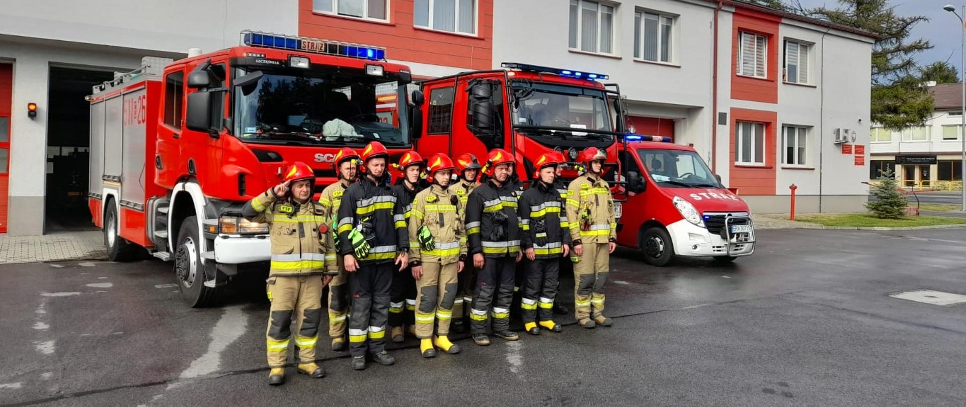Grupa strażaków w umundurowaniu specjalnym na tle samochodów strażackich. Z tyłu budynek strażnicy w kolorze szaroczerwonym.