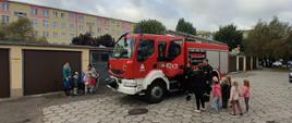Na zdjęciu widać grupę przedszkolaków oraz samochód ratowniczo-gaśniczy i strażaków. Dzieci stoją
przy pojeździe i oglądają sprzęt ratowniczo-gaśniczy. Całość odbywa się na parkingu przy przedszkolu.
W tle widać garaże oraz bloki mieszkalne.