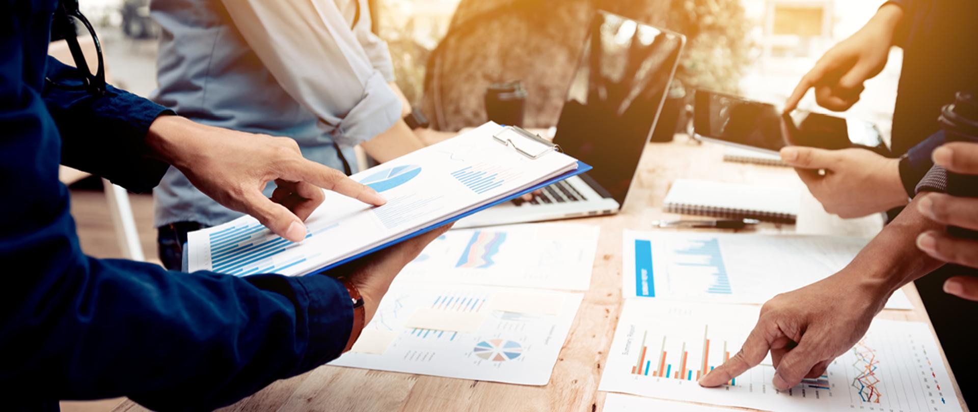 Kilka osób wskazuje palcami na dane, które są na wydruku, tablecie i komputerze 