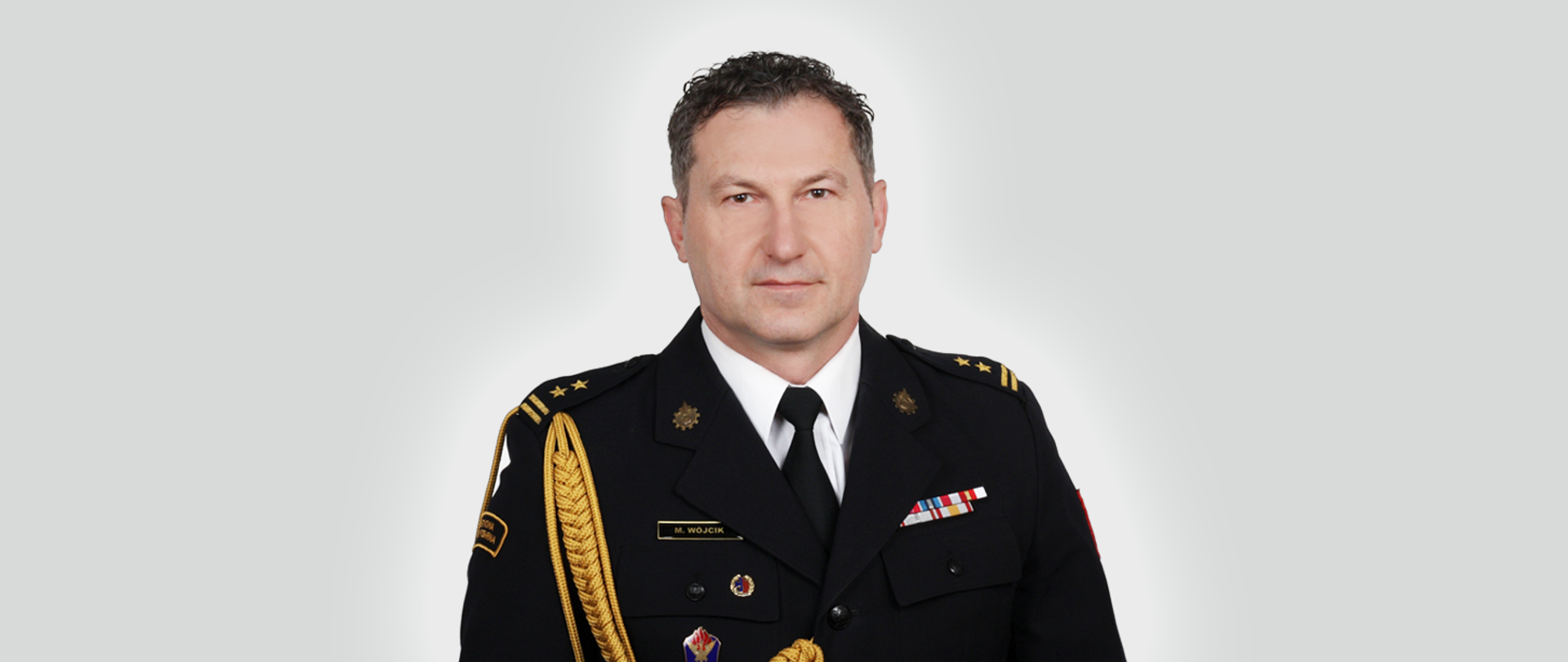 Zdjęcie przedstawia brygadiera Mariusza Wójcika Komendanta Powiatowego PSP w Świebodzinie w mundurze na szarym tle. 