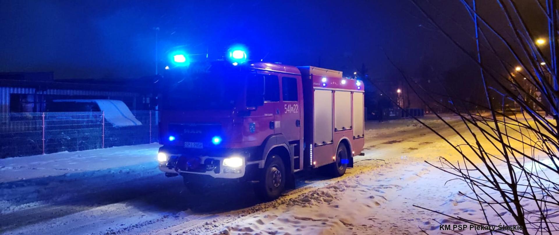 Zdjęcie przedstawia średni samochód ratowniczo - gaśniczy z włączonymi światłami ostrzegawczymi, stojący na zaśnieżonej drodze.