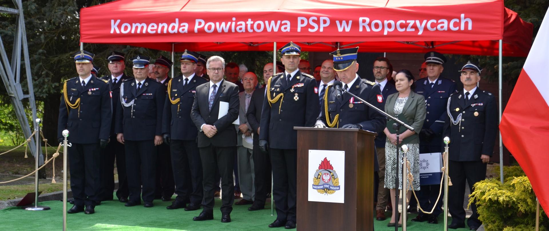 Uroczystość zrobiona na zewnątrz, podczas uroczystego apelu. Na pierwszym planie oficer Państwowej Straży Pożarnej przemawiający do mikrofonu stoi za mównicą. Za przemawiającym widać gości uroczystości stojących pod namiotem, na którym jest napis Komenda Powiatowa Państwowej Straży Pożarnej w Ropczycach.