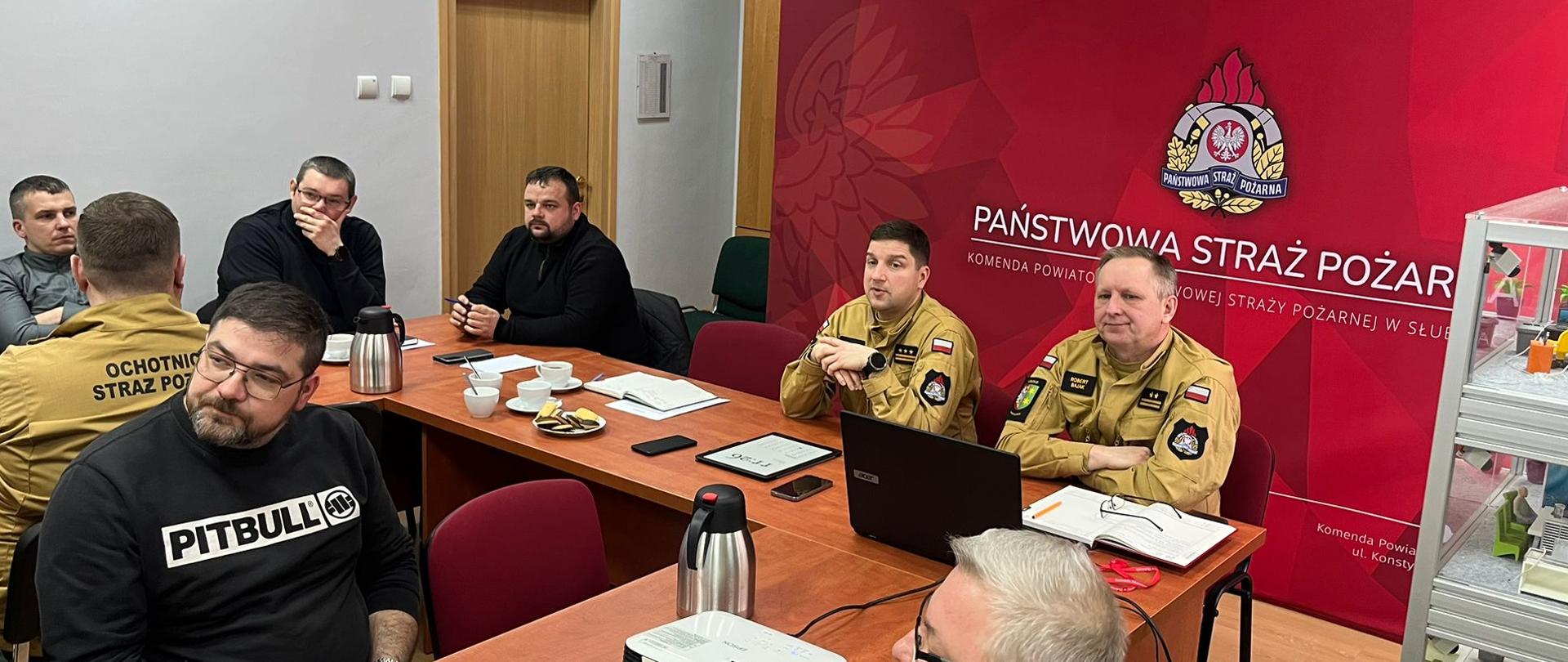 Odprawa służbowa w budynku komendy PSP, strażacy OSP ubrani po cywilnemu siedzą przy stołach naprzeciwko siebie. Komendanci w ubraniach służbowych siedzą przy biurku. 